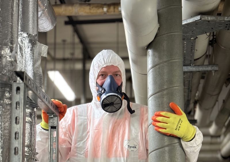 A scaffolder in a disposable coverall, hood and P3 half-face respirator working among lagged pipework in a plant room